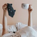 person laying in bed holding up mug and sleeping mask