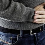Man Wearing Grey Sweater And Blue Jeans With His Hands On His Stomach Holding It Because It Hurts